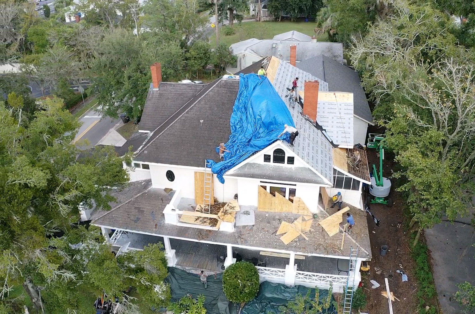 A two-story home undergoing a full tear-off and replacement of the shingle roof, with protective measures in place, like tarping and wood barriers over the windows.