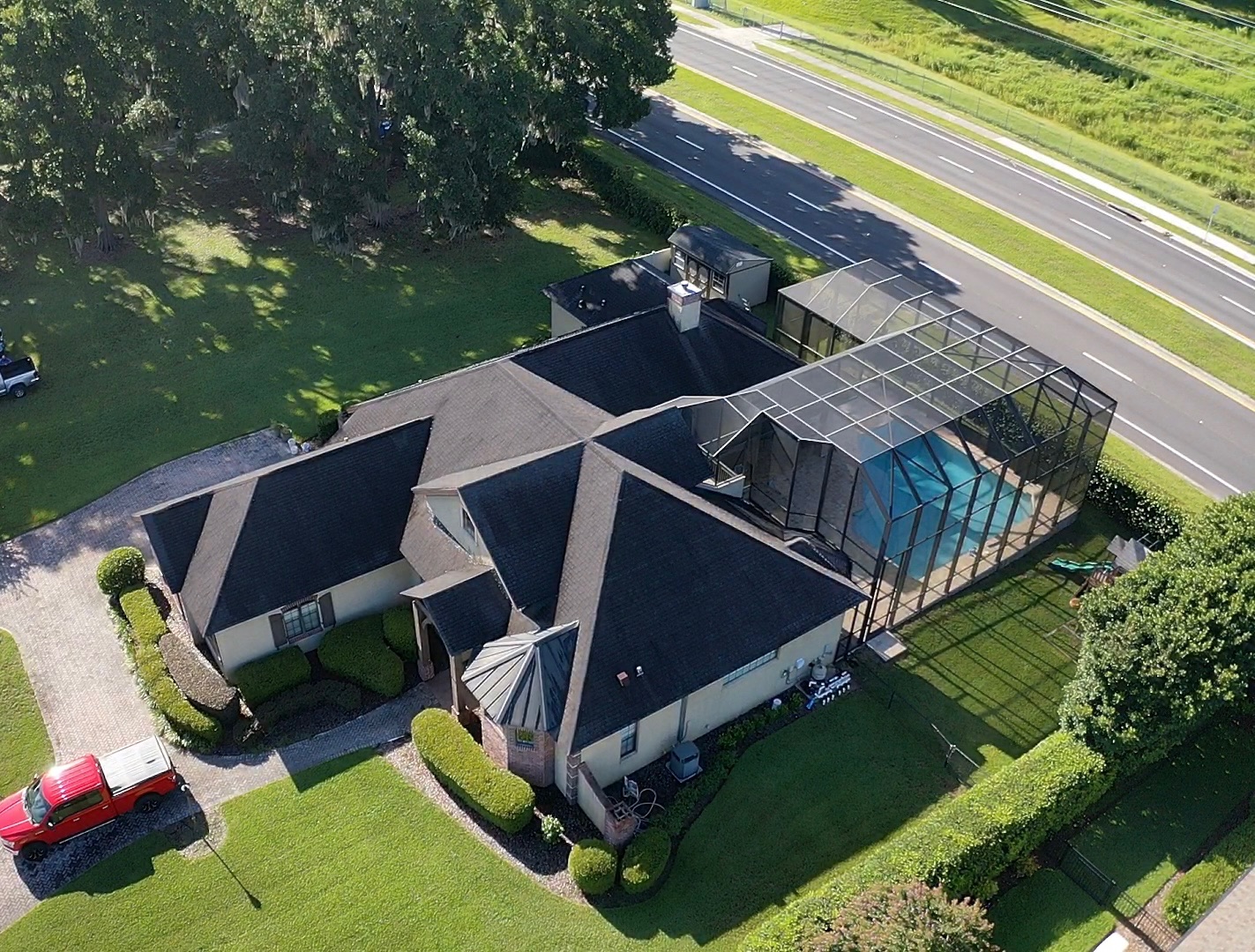 An aerial view of a stucco house with an asphalt shingle roof and a screened in pool next to a road.