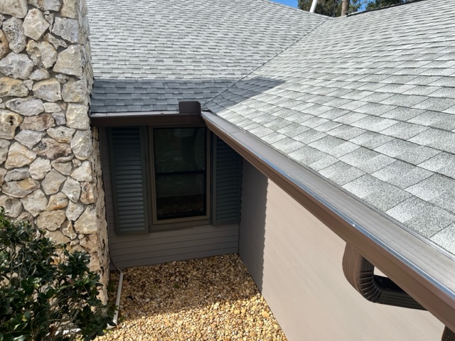A close-up view of brown gutters with gutter guards on a brown house with a stone chimney and an asphalt shingle roof.