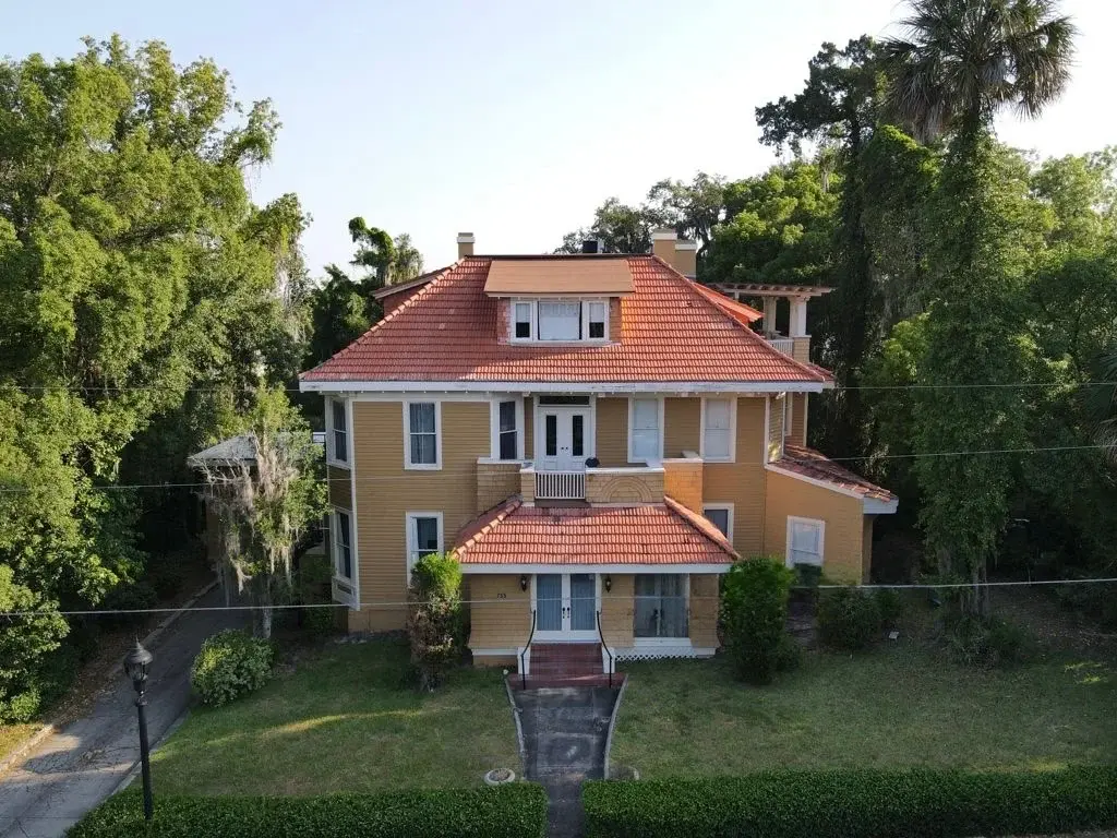 Reliable Residential Roofing In Wildwood, FL | Sentinel Roofing A three-story home with yellow siding and an orange tile roof. The house has tall, green trees on both sides, and features white gutters and trim.