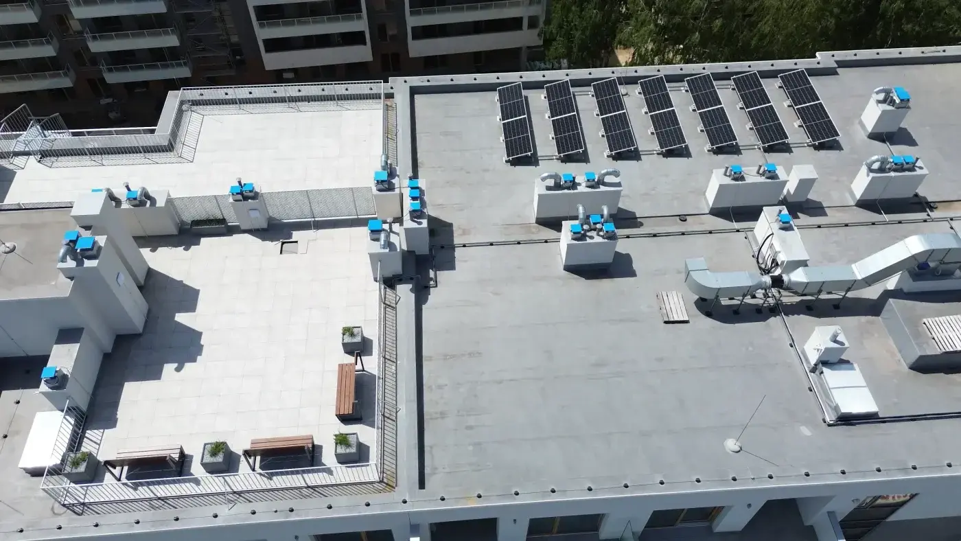 Commercial Roofing In Newberry, FL | Sentinel Roofing The roof of an apartment building has two separate sections. On the left is a living space with benches and potted plants, along with a low silver fence. The surface is white and tiled. On the right, there are solar panels, multiple HVAC units, and a light gray surface.