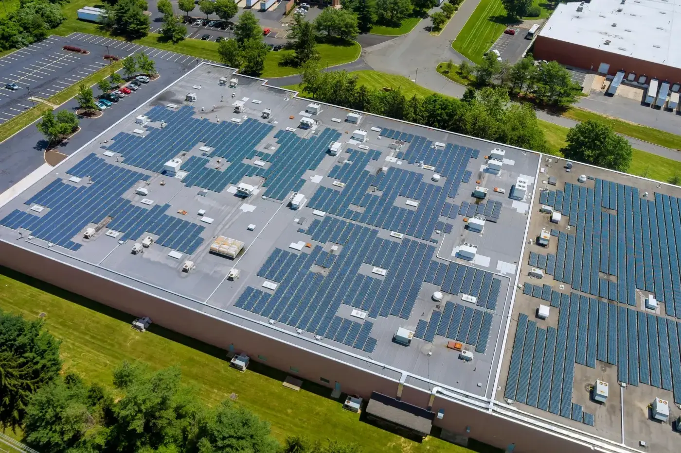 Significant Commercial Roof Repair In Oxford, FL | Sentinel Roofing An expansive warehouse with a flat roof covered in light gray material on one half, and darker gray on the other. The roof has many solar panels covering a large portion of the space. Where the panels aren’t, you can see HVAC equipment and other assorted mechanical components. The building is in an industrial parkway, with parking lots, roads, and a small grassy area visible beside it.