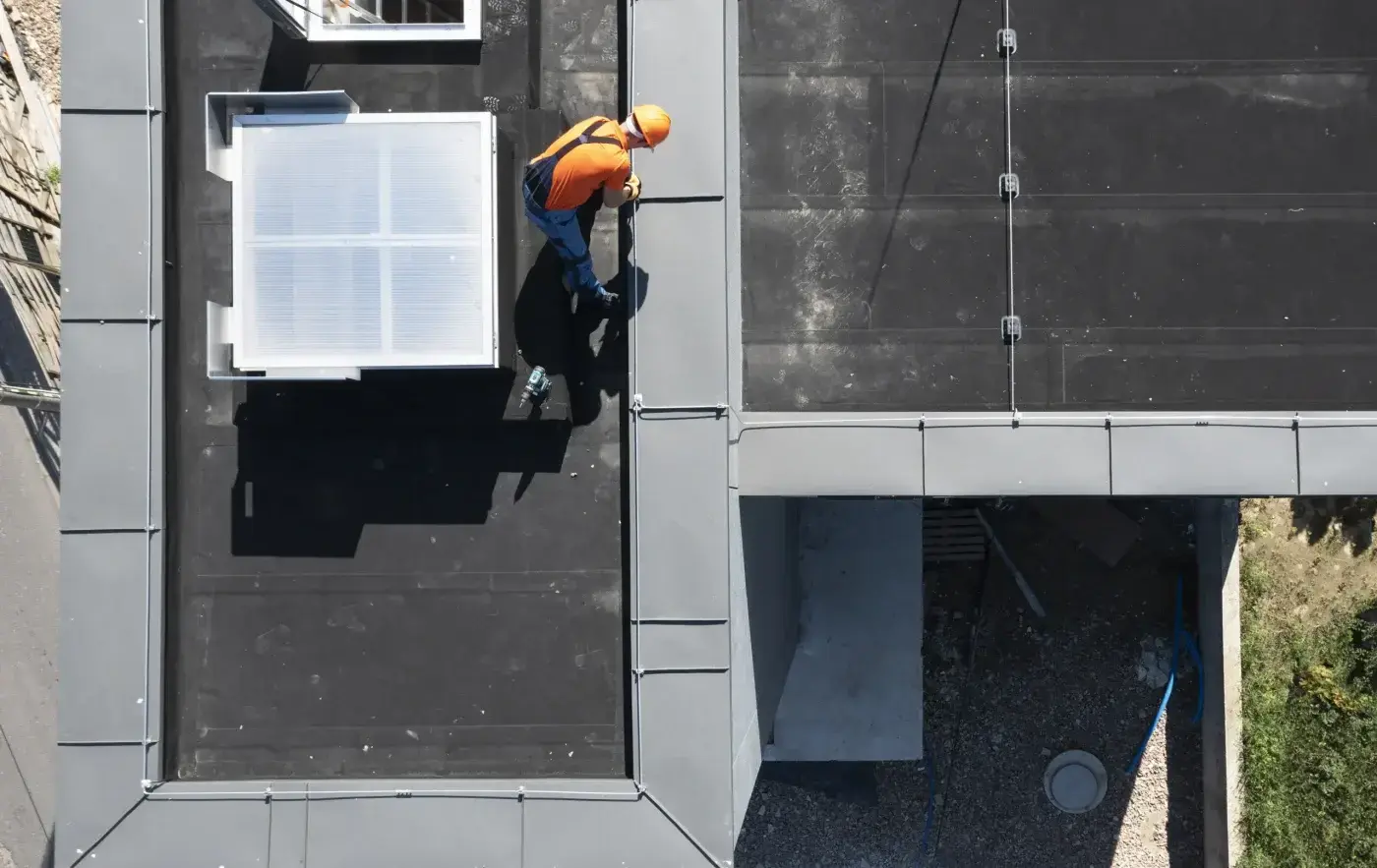 Sincere Commercial Roof Repair In Lady Lake, FL | Sentinel Roofing A man in an orange shirt and blue overalls is wearing an orange hard hat as he stands on top of a commercial roof. He’s looking down at the gray edge of the roof. The surface of the roof looks somewhat worn and is black in color.