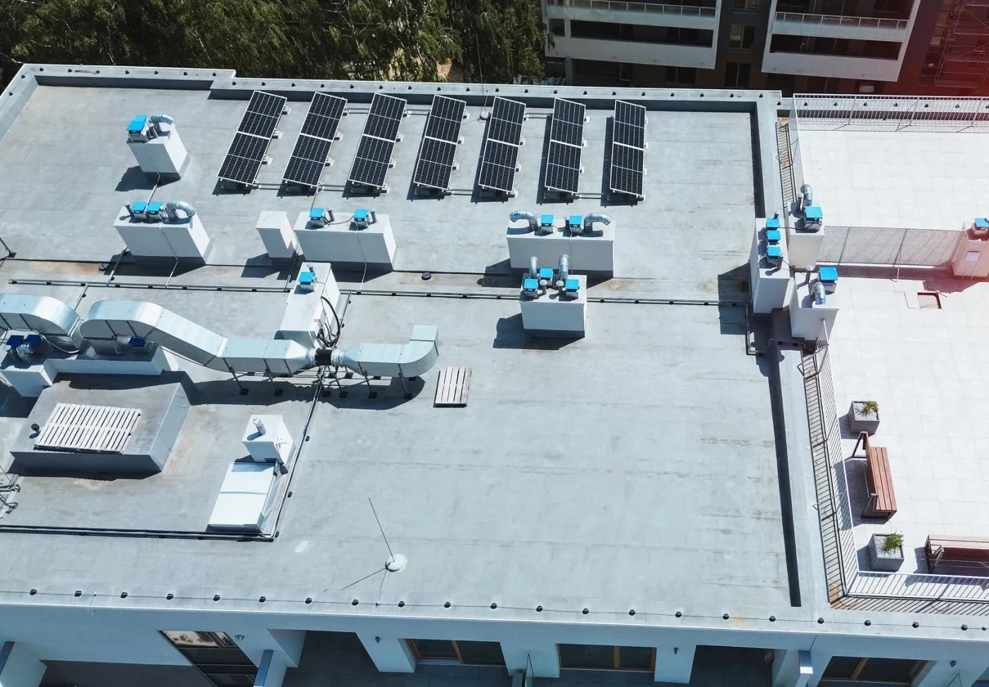Smooth-Commercial Roof Repair In Gainesville, FL | Sentinel Roofing The rooftop of an apartment complex. There are multiple HVAC unit components visible, along with solar panels. To the right, part of a sitting area with brown benches and concrete planters is visible.