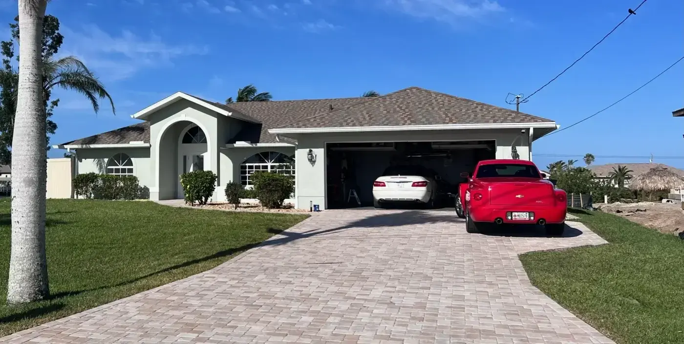 Solid Residential Roof Repair In Alachua, FL | Sentinel Roofing A single-story home with a white exterior and a large, wide driveway. The home has a dark brown shingle roof with multiple sloped sections. There are two vehicles parked in the driveway, a red one and a white one.
