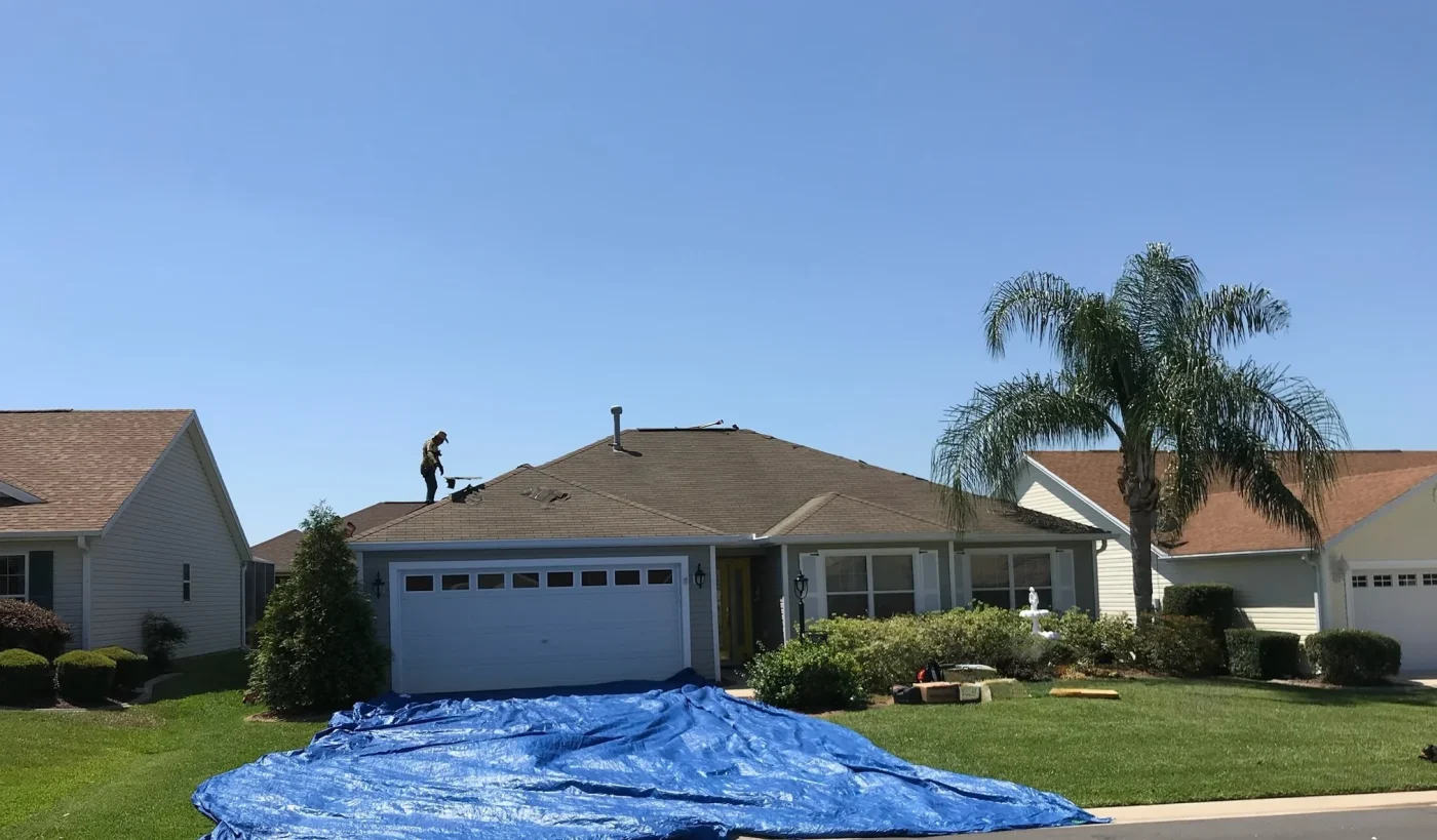 Steady Residential Roof Repair In Newberry, FL | Sentinel Roofing A man is standing on the brown-shingled roof of a home with a double-wide garage door. There is a blue tarp laid down and covering the entire driveway.
