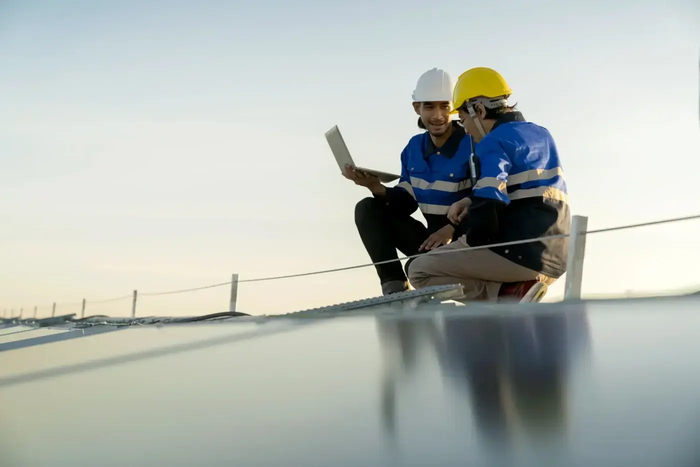 Thorough Commercial Roof Repair In Lady Lake, FL | Sentinel Roofing Two men wearing protective gear are crouched down on top of a slightly sloped commercial roof to look at a laptop. The men are wearing blue and white jackets and hard hats. The roof’s surface is dark colored, and there is a low rope barrier just in front of them.